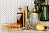 A bar setup with a bottle of Ghia, a whiskey box, a gold cocktail shaker, four glasses, a large yellow lemon, and green plant against a herringbone tile backsplash.