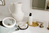 A bathroom counter with a white pitcher, round mirror on a green towel, speckled soap dish, lotion bottle on a gold tray, and a faucet.