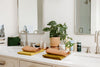 A bathroom counter with dual sinks, two mirrors, a potted plant, toiletries, and neatly folded towels.