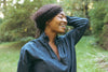 A person wearing a denim shirt smiles with their eyes closed, standing outdoors with greenery in the background.