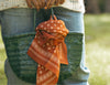 Person wearing a knit sweater and jeans holds a green woven bag with a red patterned scarf tied to it, standing outdoors.