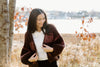 A woman wearing a striped cardigan stands near trees and a body of water, smiling and looking down. Autumn leaves surround her.