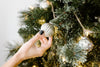 A hand places a silver ornament on a decorated Christmas tree with white lights and green branches.