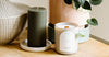 Green ribbed candle and white scented candle on a wooden surface, next to potted plants.