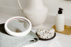 Bathroom counter with a round mirror, knitted cloth, speckled soap on a dish, and a lotion bottle on a golden tray.