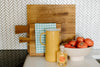 A yellow pitcher, hand soap, and a bowl of tomatoes are on a countertop. Two wooden cutting boards with a checkered cloth lean against a white, textured tile wall.