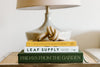 A lamp sits on a table behind three stacked books. Books are titled 