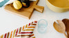 A wooden cutting board with a lemon and a dark jar, a striped cloth, a yellow bowl, a glass of water, and wooden utensils on a white surface.