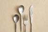 A set of four rustic silver utensils on a light wood surface: two spoons, one fork, and one knife.