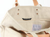 Open beige canvas tote bag with tan leather handles, displaying a label that reads 