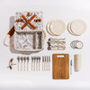 Picnic set with an open basket, plates, cups, cutlery, corkscrew, and a wooden cutting board arranged neatly on a white surface.