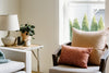 A cozy living room corner with a chair, beige and orange pillows, a side table with a lamp, candles, plant, and light streaming through a window.