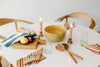 A round white table with a yellow bowl, wooden utensils, striped napkins, glasses, candles, and a cutting board with a lemon and a bottle.