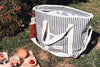 A large striped tote bag sits on grass beside a wooden board with sliced citrus fruits and two glasses of iced beverages.