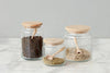 Three glass jars with wooden lids and spoons, filled with peppercorns, dried herbs, and sunflower seeds, are placed on a marble surface.