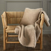 A wooden chair with woven seat and backrest is draped with a soft, beige blanket and pillow, set against a gray wall on a wooden floor.