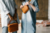 Two people stand in denim outfits; one holds a brown leather bag with a chain handle, and the other adjusts a brown leather crossbody bag.