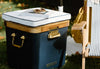 A cooler with a cushion top is used as a side table with a book and sunglasses on it, beside a striped chair and a yellow bag on the grass.