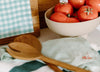 A bowl of ripe tomatoes sits on a kitchen counter next to wooden spoons, a folded tea towel, and a checkered cloth-covered board.