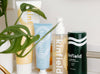 A collection of Kinfield skincare products displayed on a shelf, partially obscured by a green plant leaf.