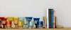 Colorful glassware and books are neatly arranged on a wooden shelf, with stone bookends holding the books upright against a white background.