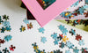 Puzzle pieces scattered on a white surface with two open puzzle boxes, one pink and one green, visible in the background.