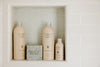 Four Flamingo Estate bath products, including body wash, lotion, body oil, and soap, are displayed on a tiled shelf.