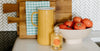 A yellow pitcher, a bottle, a bowl of tomatoes, and a wooden cutting board with a checkered cloth, are arranged on a kitchen countertop.