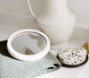 A round mirror rests on a textured gray fabric next to a white vase and a spotted dish on a white surface.