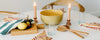 A round table set with a yellow bowl, salad utensils, striped napkins, lemons, olive oil, two glass mugs, and three lit candles.