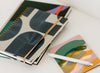 A stack of notebooks with geometric patterns and a rubber band, next to a pen and a colorful notepad, on a white surface.