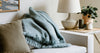 A cozy living room scene with a white sofa, draped with a fluffy blue blanket and a gray pillow. A wooden side table holds a textured lamp, candles, and a potted plant.