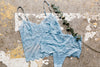 A blue lace camisole and panty set with thin straps is laid out flat on a textured surface, accompanied by a sprig of eucalyptus.