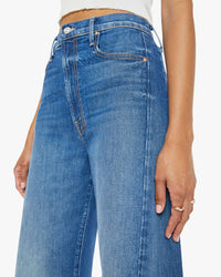 A person wears high-waisted, Mother Full Pipe Flood jeans in It's A Small World denim blue with a white top, shown from the waist down. One hand rests near the pocket.