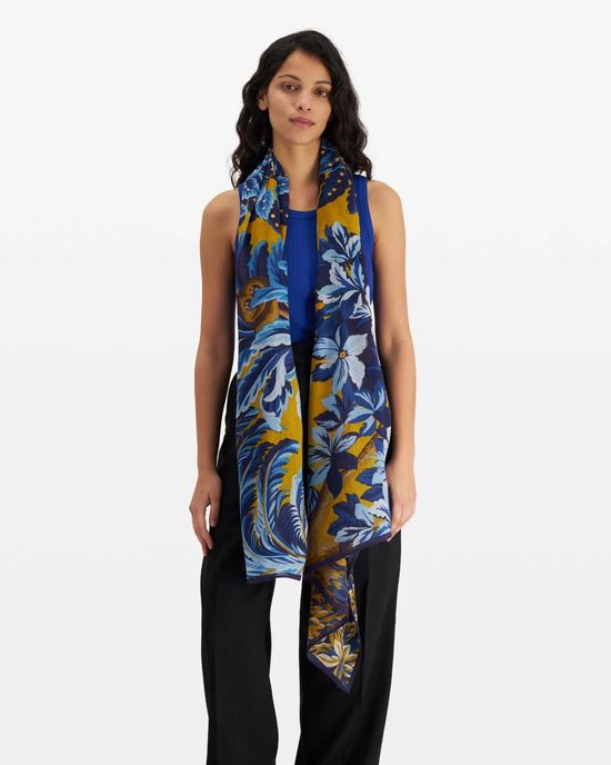 A woman modeling the Etole 100 Chatou Winter Scarf in Duck Blue by Inoui Editions, a 100% Wool, blue and gold floral scarf draped over a plain top and black trousers.