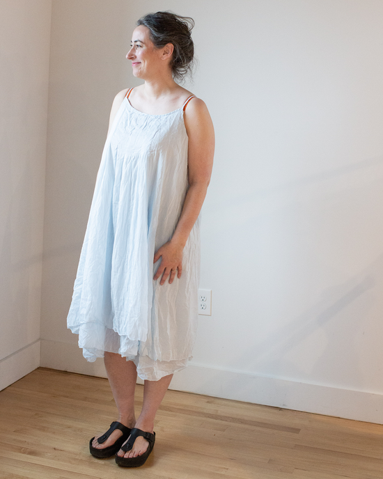A woman in a CP Shades Lia Darted Front Dress in Seafoam Cotton Silk and sandals standing indoors, smiling and looking to the side.