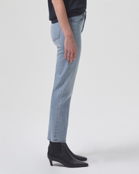Person standing side-on wearing AGOLDE Kye in Diversion mid-rise straight leg Cake Washed blue jeans and black boots against a white background.