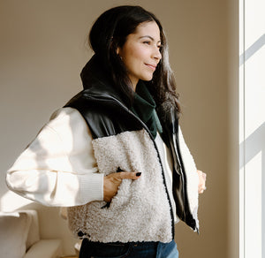 A woman wearing a white sherpa and black leather jacket stands in a sunlit room, hands in pockets, looking to the side.