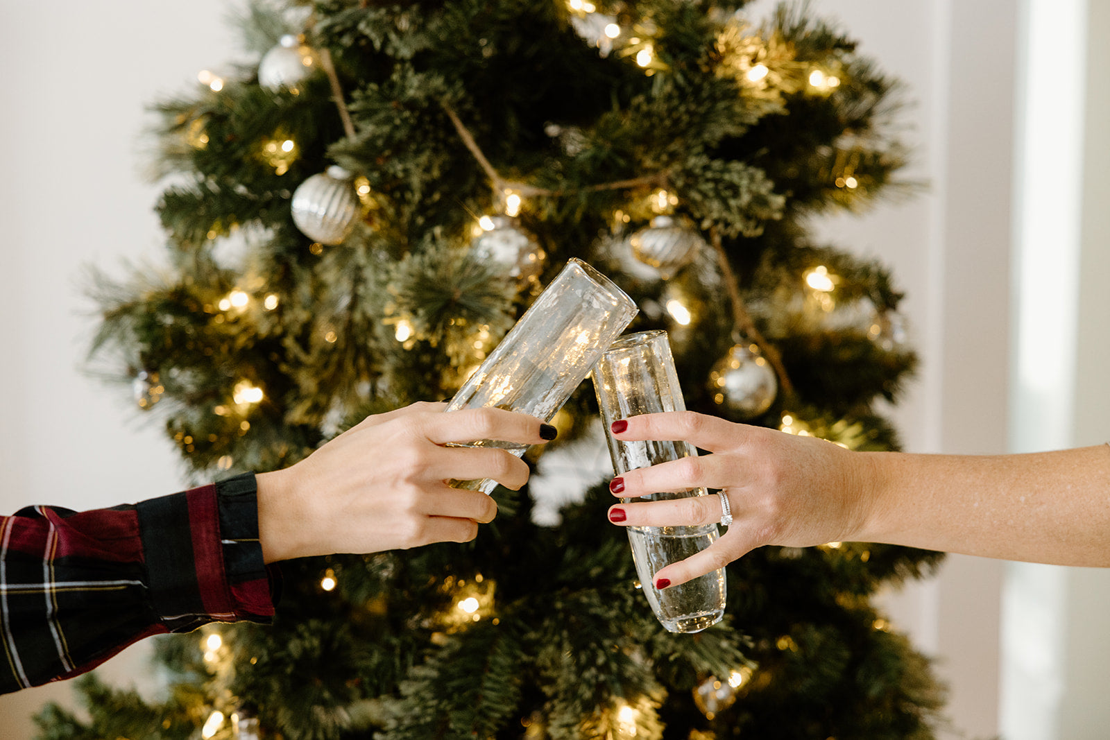 Two hands clink champagne glasses in front of a decorated Christmas tree with lights and ornaments.