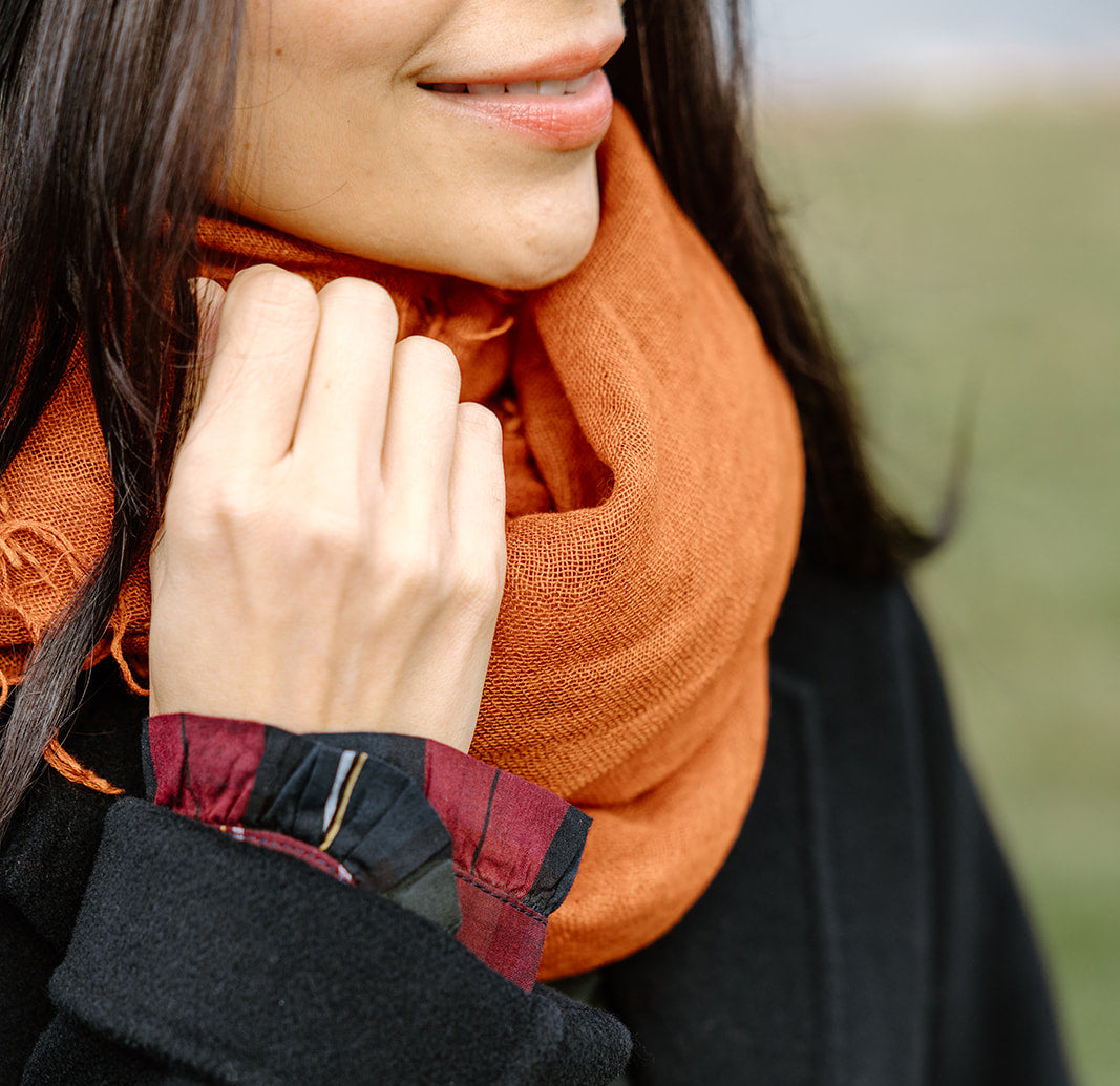 A person wears an orange scarf and a black coat, holding the scarf. The background is blurred.