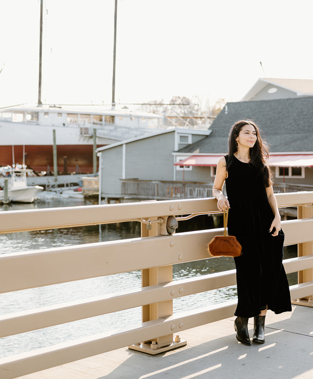 A woman in a black dress walks on a bridge near a harbor with boats and buildings in the background.