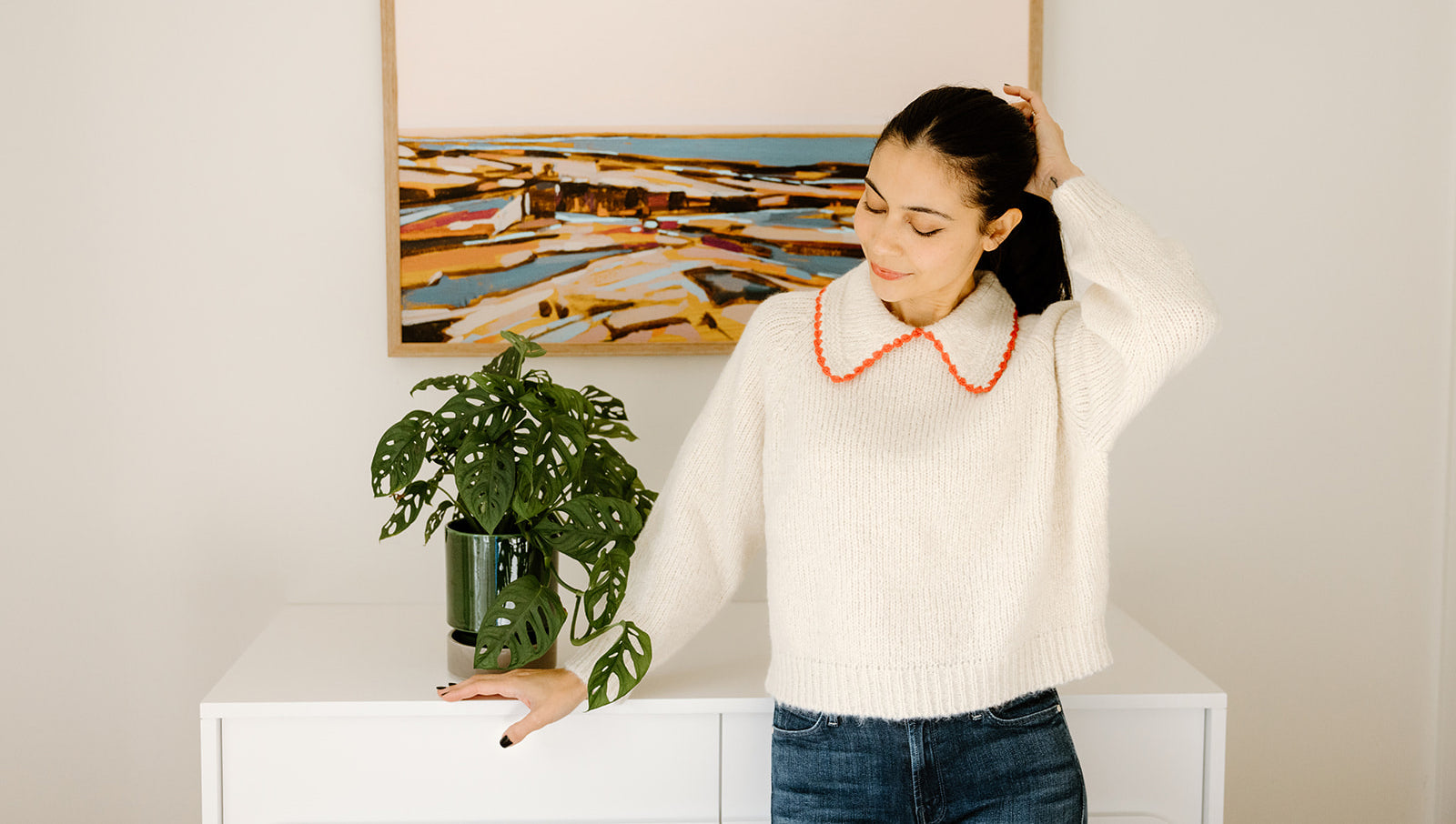 A person stands next to a plant, wearing a white sweater and red necklace. A colorful landscape painting hangs on the wall behind them.