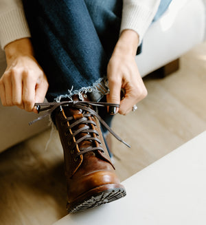 Person tying the laces of a brown leather boot while sitting, wearing blue jeans and a cream sweater.