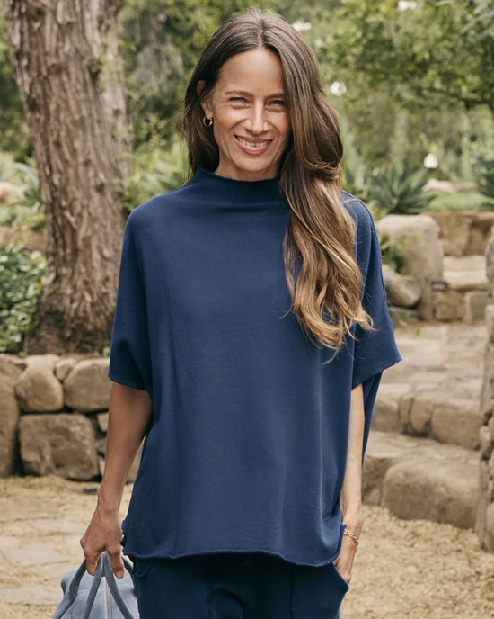 A smiling woman wearing a blue Audrey Funnel Neck Capelet in Air Force by Frank & Eileen standing outdoors.