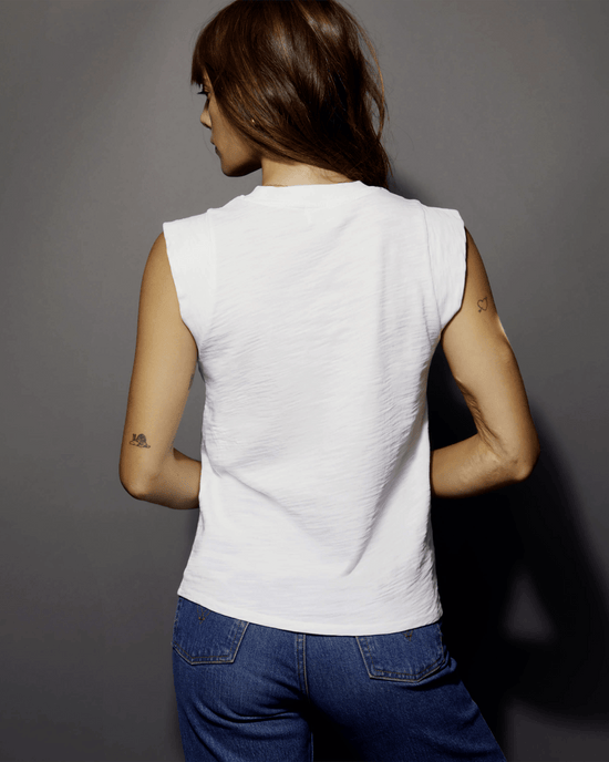 A person with long hair wearing a Patti Muscle Tank in Optic White by Nation LTD and blue jeans stands facing away, displaying small tattoos on both arms.