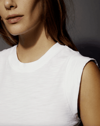 Close-up of a person wearing the Patti Muscle Tank in Optic White by Nation LTD against a plain background. Their face is partially visible, showing part of the nose, mouth, and chin. Their hair is brown, and their left shoulder is visible.