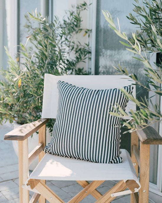 A Small Square Throw Pillow in Laurens Navy Stripe by Business & Pleasure Co adorns a wooden director's chair made from UV-durable Pleasuretex fabric, set elegantly amid potted green plants in front of a building.