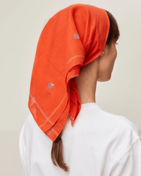 A person with long hair is wearing Moismont's Bandana No 656 in Poppy Red, a handcrafted Jamdani headscarf featuring small decorative shapes, viewed from the side and back against a plain background.