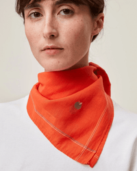 A person wearing a white shirt and Moismont's Bandana No 656 in Poppy Red, an orange handcrafted cotton bandana with a small embroidered logo, around their neck.