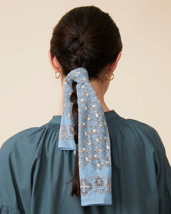 A woman with a braid tied with a Moismont Bandana No 675 in Cloud, wearing a teal blouse and gold hoop earrings, faces away.